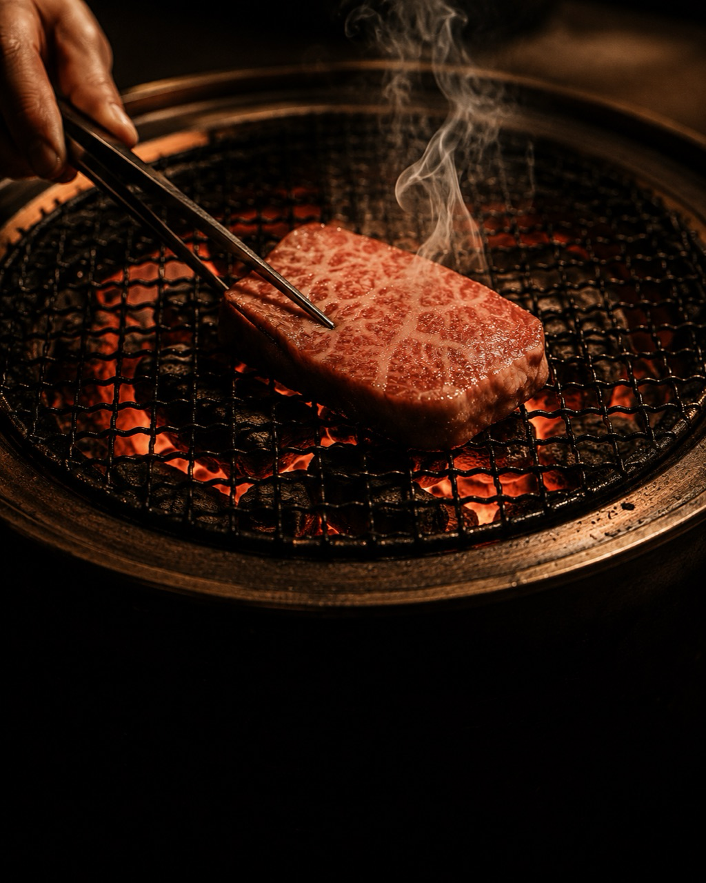 七輪の備長炭の上で和牛をトングで掴む店主の手元、立ちのぼる煙